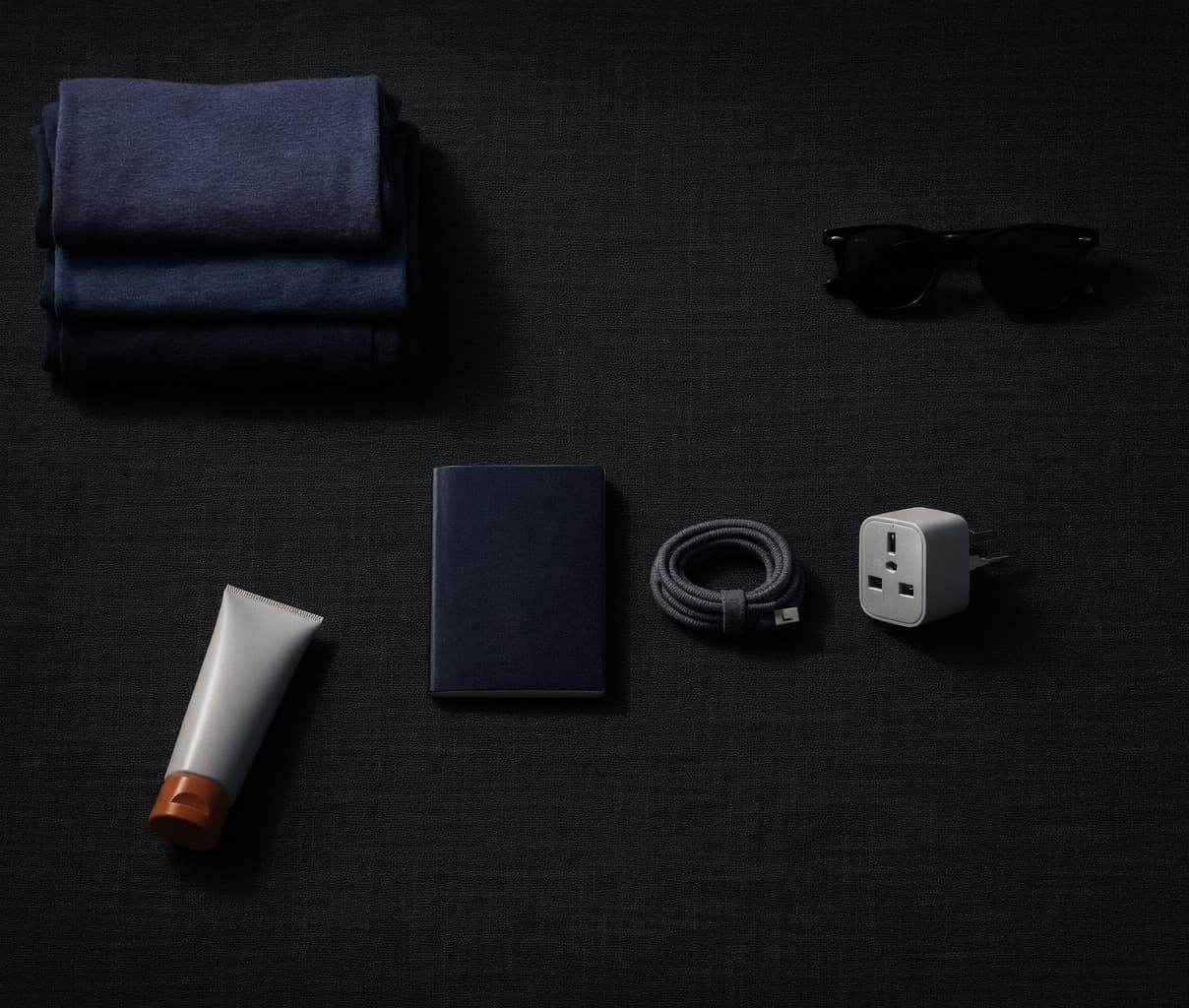 Travel clothes, sunglasses, an adapter and sunscreen laid out beside a bag.