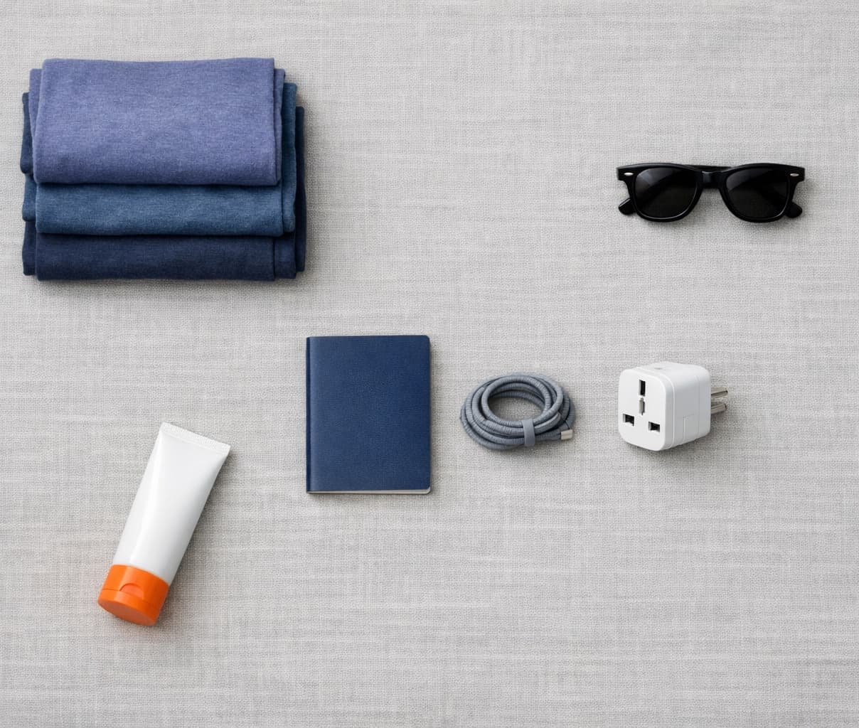 Travel clothes, sunglasses, an adapter and sunscreen laid out beside a bag.
